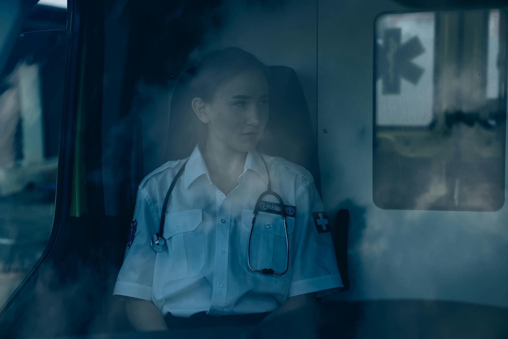 A thoughtful paramedic sits inside an ambulance, wearing a uniform and stethoscope.